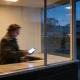 A person in a coat paying with a phone at a counter seen from outside a glowing shop window at dusk, their reflection on the glass and the cold blue street beyond