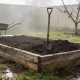 An empty raised garden bed of weathered hardwood sleepers holding heaped dark soil, a spade standing in it and a wheelbarrow beside, on wet lawn in a misty Australian backyard