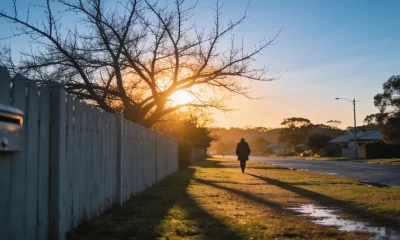 A quiet Australian suburban street at sunset in mid-winter, bare branches in front of a low ochre sun, long shadows on wet grass and a person in a coat walking away in the distance