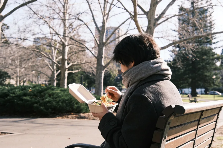 A person in a coat and scarf eating lunch on a park bench in pale winter sun, bare trees and evergreen shrubs in an Australian city park behind.