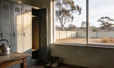 A depot smoko room at the end of a shift, a stainless steel kettle on a worn timber table, an open grey locker with work boots below and a high window onto a dry Australian yard