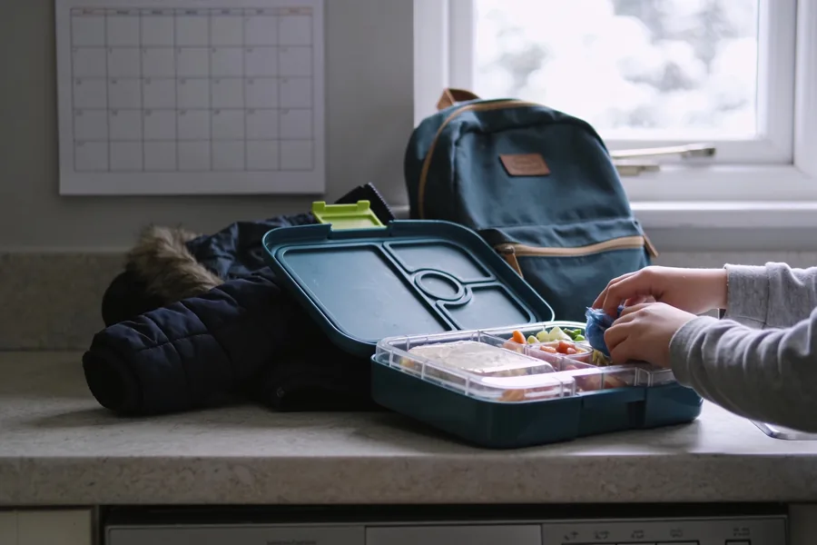 A child's lunchbox being packed on a kitchen bench in winter daylight, a backpack, a coat and a wall calendar beside it, hands only in frame.