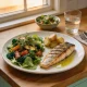 A home meal on a timber table: grilled fish, roast potatoes and a large salad on a plate with olive oil, a glass of water and cutlery on a napkin