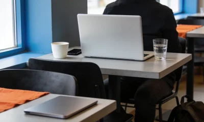A person seen from behind working on a laptop at a cafe table, its screen facing away, with a coffee, a glass of water and a phone beside it