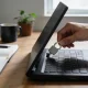 A hand holding a small security key towards a laptop's side port on a timber desk, with a notebook of blank pages, a pen, a mug and a plant beside it