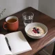 A kitchen table with a blank notebook and pen beside a breakfast of porridge with berries, a mug of tea, water and a potted herb