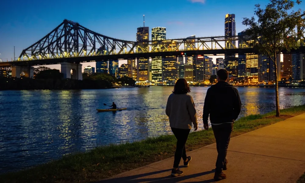 Two people walking away along the Brisbane riverwalk at dusk with the lit Story Bridge, the city skyline and a kayaker on the river