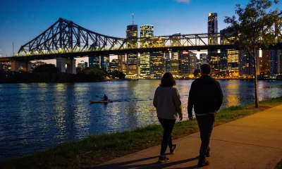 Two people walking away along the Brisbane riverwalk at dusk with the lit Story Bridge, the city skyline and a kayaker on the river