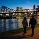 Two people walking away along the Brisbane riverwalk at dusk with the lit Story Bridge, the city skyline and a kayaker on the river