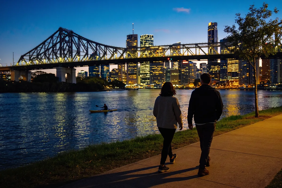 Two people walking away along the Brisbane riverwalk at dusk with the lit Story Bridge, the city skyline and a kayaker on the river