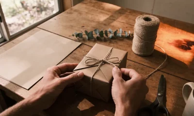 Hands tying twine around a brown-paper-wrapped box on a timber table, with a spool of twine, eucalyptus sprig, plain paper and scissors