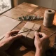 Hands tying twine around a brown-paper-wrapped box on a timber table, with a spool of twine, eucalyptus sprig, plain paper and scissors