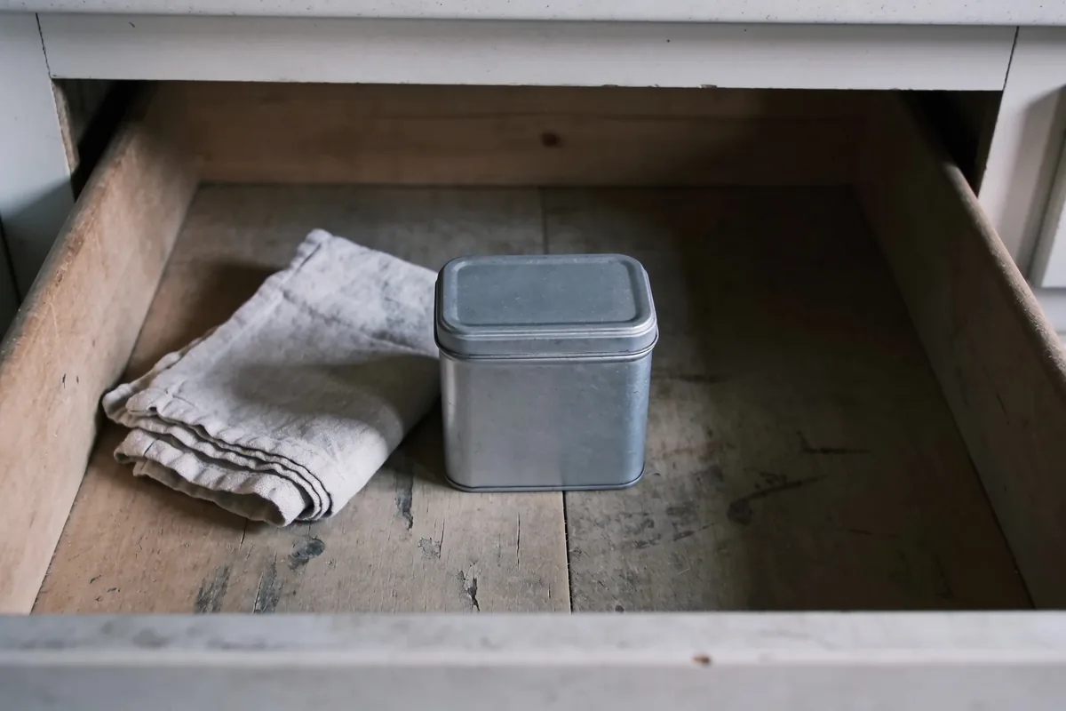 An open kitchen drawer in an Australian home holding a plain lidded tin and a folded cloth, in soft winter morning light