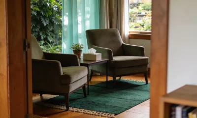 Two armchairs facing each other across a low table with a plant and a tissue box in a calm counselling room, daylight through curtains