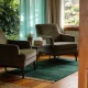 Two armchairs facing each other across a low table with a plant and a tissue box in a calm counselling room, daylight through curtains