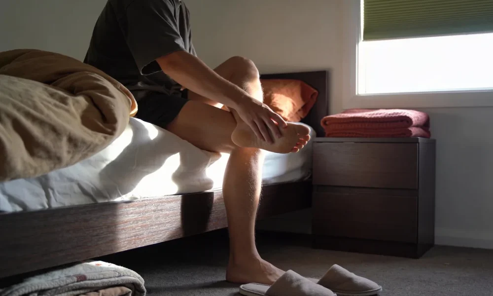A person sitting on the edge of a bed in morning light, one foot lifted onto the opposite knee with a hand pressed along the arch and heel, slippers and folded bedding nearby