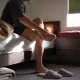 A person sitting on the edge of a bed in morning light, one foot lifted onto the opposite knee with a hand pressed along the arch and heel, slippers and folded bedding nearby
