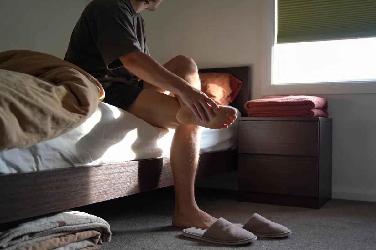 A person sitting on the edge of a bed in morning light, one foot lifted onto the opposite knee with a hand pressed along the arch and heel, slippers and folded bedding nearby