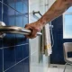 An older adult's hand gripping a steel grab rail on a tiled shower wall, with a folded towel and a shower chair beside it in daylight