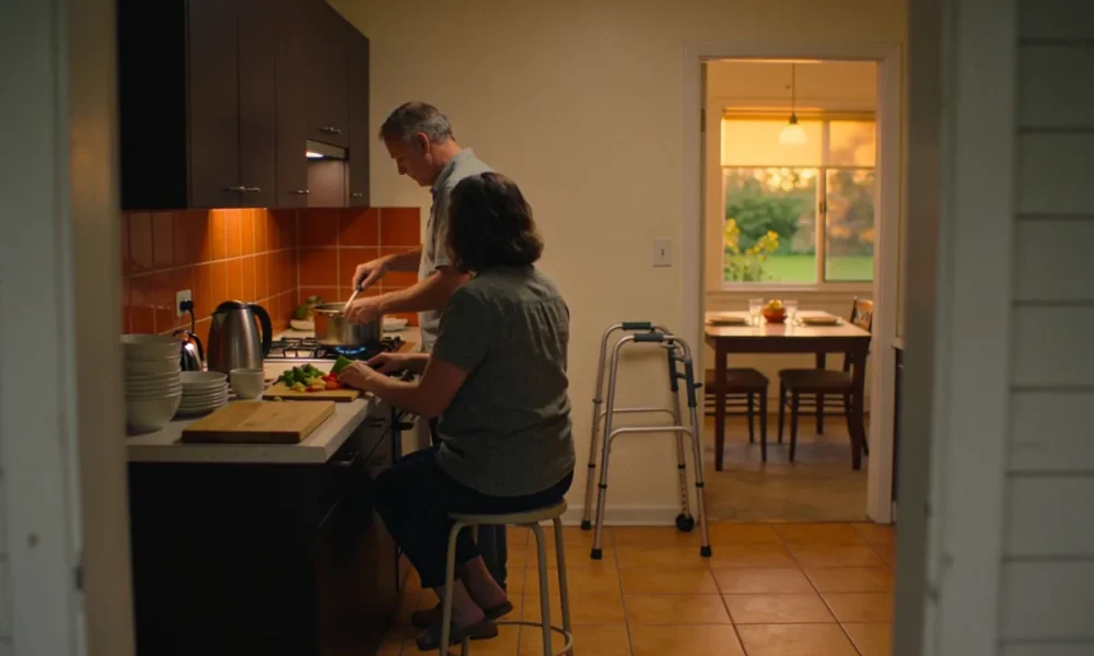 Two adults cooking an evening meal together in a home kitchen, one seated at the bench, with a folded walking frame against the wall and a table set beyond
