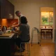 Two adults cooking an evening meal together in a home kitchen, one seated at the bench, with a folded walking frame against the wall and a table set beyond