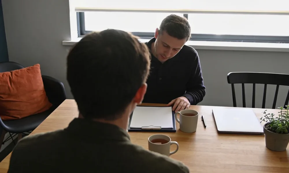 Two people in conversation across a timber table, a clipboard of blank paper between them, mugs, a closed laptop and a plant, daylight from a window