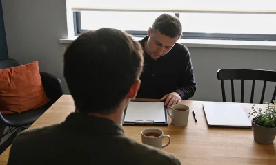 Two people in conversation across a timber table, a clipboard of blank paper between them, mugs, a closed laptop and a plant, daylight from a window