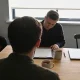 Two people in conversation across a timber table, a clipboard of blank paper between them, mugs, a closed laptop and a plant, daylight from a window