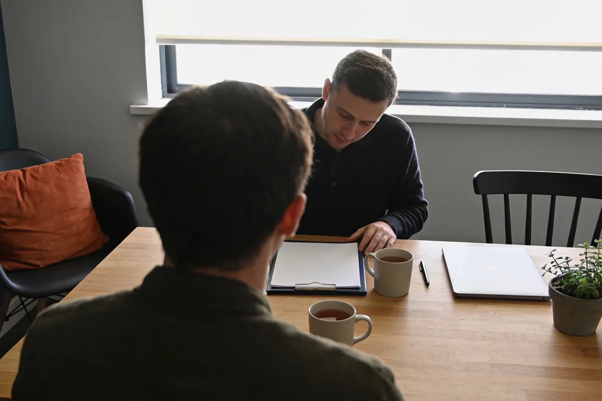 Two people in conversation across a timber table, a clipboard of blank paper between them, mugs, a closed laptop and a plant, daylight from a window