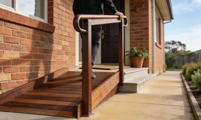 A timber access ramp with a handrail leading to the front door of a brick home, a person walking up with one hand on the rail, garden path beside it