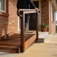 A timber access ramp with a handrail leading to the front door of a brick home, a person walking up with one hand on the rail, garden path beside it