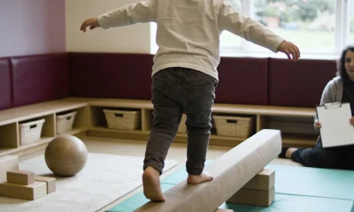 A young child seen from behind balancing along a low beam in a bright playroom, with an adult seated nearby holding a clipboard of blank paper