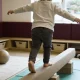 A young child seen from behind balancing along a low beam in a bright playroom, with an adult seated nearby holding a clipboard of blank paper