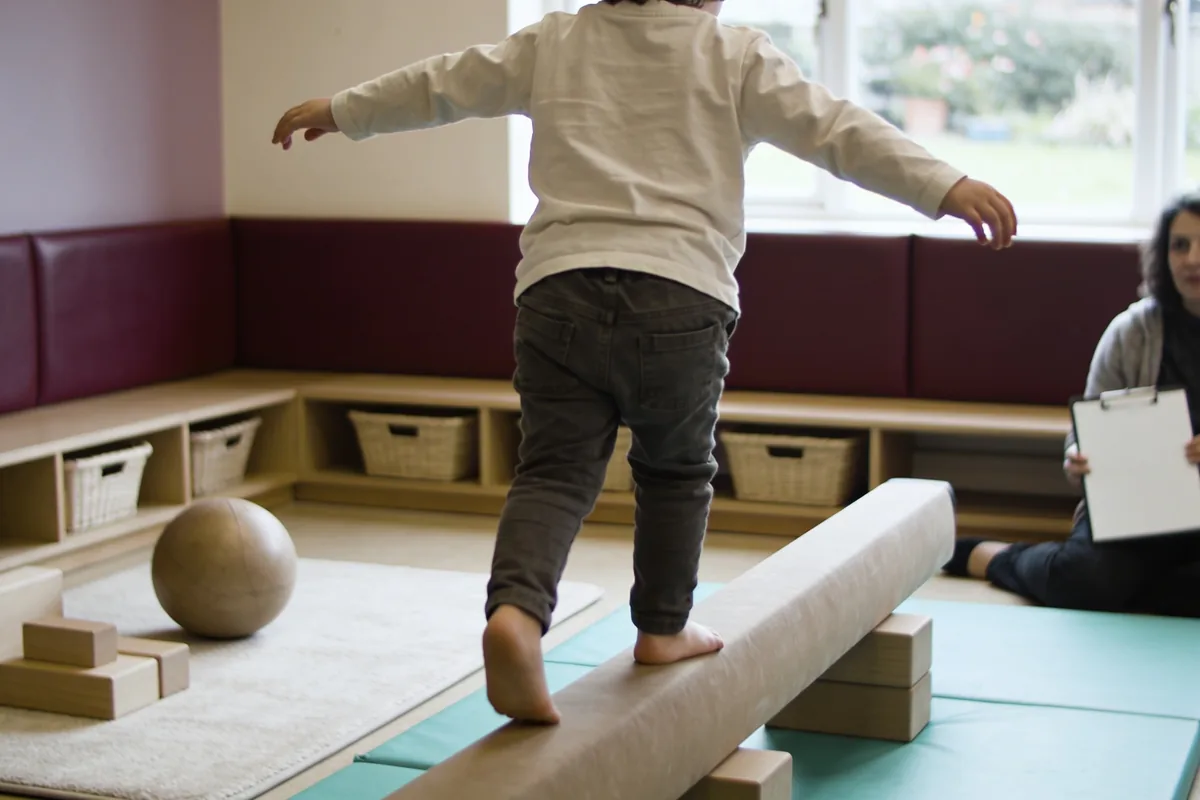 A young child seen from behind balancing along a low beam in a bright playroom, with an adult seated nearby holding a clipboard of blank paper