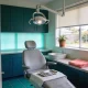 A dental surgery seen from the doorway, with the chair, overhead light and a tray of instruments on a towel, a basin and a garden window behind