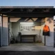 A small suburban workshop unit open at dawn with tools on a pegboard, a workbench, a trolley and a broom inside, and a high-visibility jacket on a hook