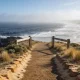 A clifftop coastal walk closed by a plain timber and mesh gate, with the ocean breaking below and a long beach curving away under morning haze