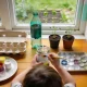 A small child seen from behind painting a recycled jar at a kitchen table, with an egg carton, cardboard tube, paint pots, bottle caps and seedlings around them