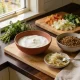A kitchen bench with a bowl of yoghurt, a bowl of lentils, a dish of sauerkraut, a board of chopped vegetables and fresh herbs
