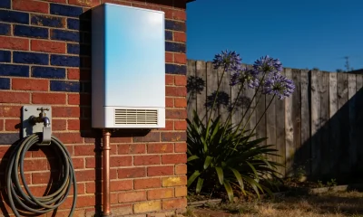 A white tankless hot water unit mounted on the exterior brick wall of a suburban home, with a copper pipe, garden tap and coiled hose in morning light
