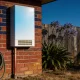 A white tankless hot water unit mounted on the exterior brick wall of a suburban home, with a copper pipe, garden tap and coiled hose in morning light
