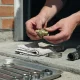 Hands holding a brass cooling-system fitting over a folded cloth on a concrete driveway, with a hose clamp, a radiator hose and a tray of spanners beside them
