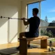 A person seated on a bench pulling a resistance band anchored to a wall, seen from behind in a sunlit room with a garden through the window