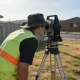 A surveyor in a high-visibility vest sighting through a total station on a tripod on a suburban house block, timber boundary post and fence behind