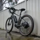 An unbranded e-bike leaning against a paling fence in a suburban Australian yard, its hub motor, coiled cable over the handlebar and disc brake visible on wet concrete