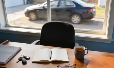 A home desk with an open notebook of blank pages, a pen, car keys, glasses and a mug of tea, with a car parked in the driveway beyond the window