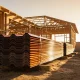 Stacks of new corrugated steel roofing sheets on timber bearers at a suburban building site, with the timber frame of a new house behind in late-afternoon sun