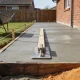 A freshly poured concrete driveway held in timber formwork with a screed board and float on the surface, and a wheelbarrow and shovel on the lawn beside it