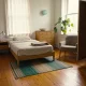 A neat bedroom in a shared home with a made bed, a chest of drawers, an armchair by the window with a plant, and an open wardrobe with hanging clothes