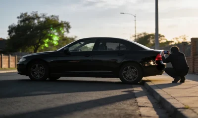 A person crouched beside a dark prestige sedan at the kerb photographing its paintwork with a phone, backlit in late-afternoon light